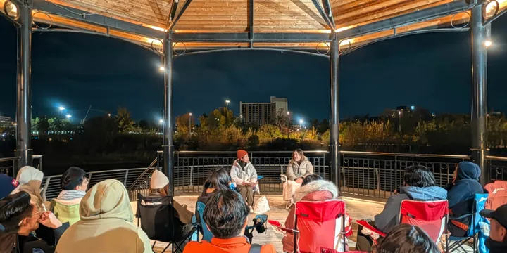 À noite, o público se reúne no Music Pavilion, aberto nas laterais, em Calgary; ao centro, duas pessoas sentadas compartilham histórias.